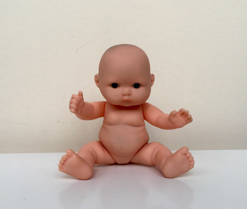 May include: A small, nude-colored baby doll with black eyes and a realistic face. The doll is sitting with its legs crossed and its arms outstretched.