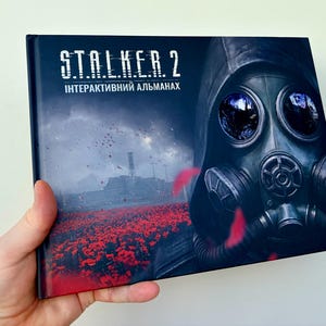 Könnte beinhalten: Ein gebundenes Buch mit dem Titel STALKER 2 ІНТЕРАКТИВНИЙ АЛЬМАНАХ, das ein dunkles Cover mit einer Gasmaske, einem Feld aus roten Mohnblumen und einem Industriegebäude unter einem bewölkten Himmel mit schwebenden roten Blütenblättern zeigt.