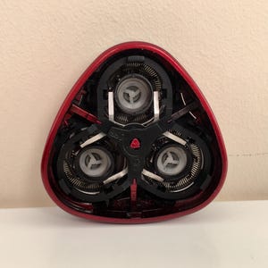 May include: Close-up of a red electric razor head with three rotating blades. The blades are visible through the clear plastic housing.