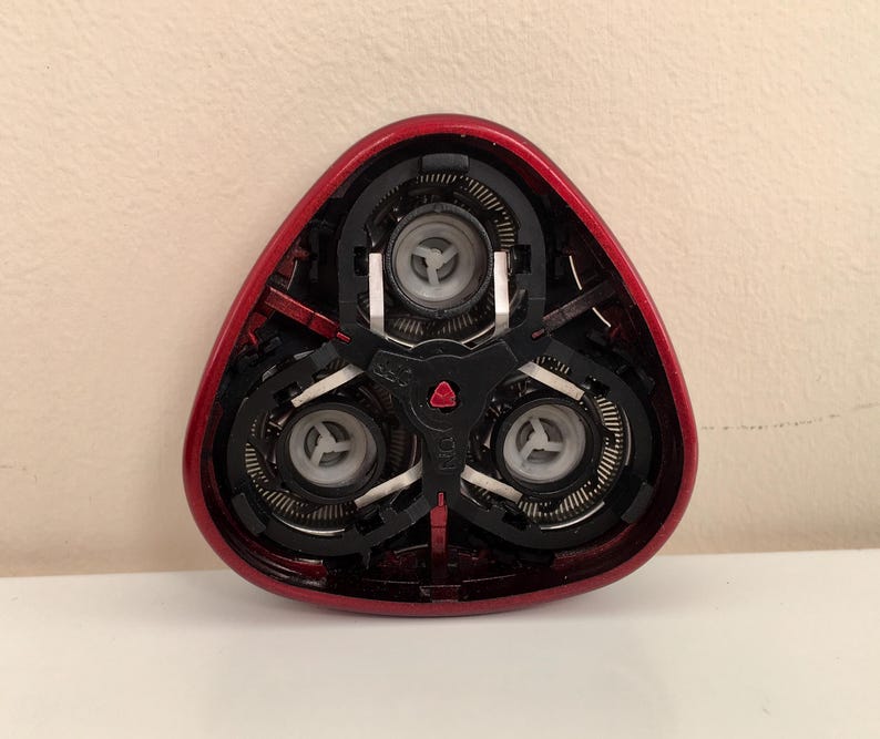 May include: Close-up of a red electric razor head with three rotating blades. The blades are visible through the clear plastic housing.