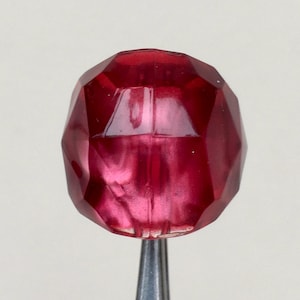 May include: A faceted, deep red glass bead with a metallic sheen. The bead is oval shaped and has a smooth, polished surface.