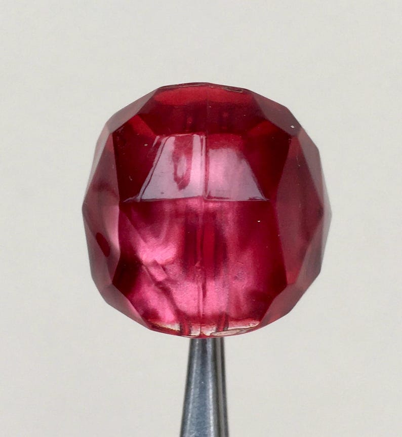 May include: A faceted, deep red glass bead with a metallic sheen. The bead is oval shaped and has a smooth, polished surface.