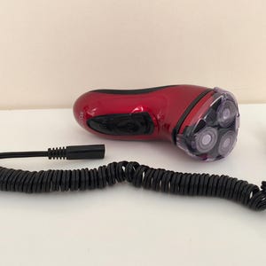 May include: A red electric razor with a coiled cord and a plug. The razor has three rotating blades.