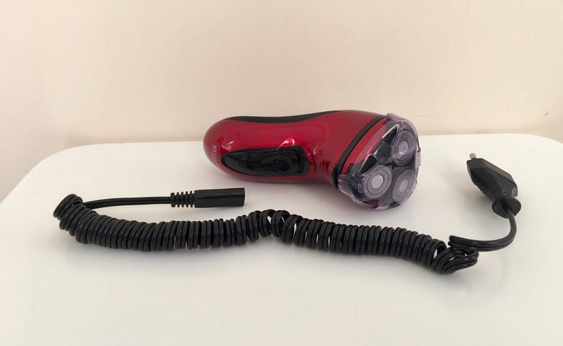May include: A red electric razor with a coiled cord and a plug. The razor has three rotating blades.