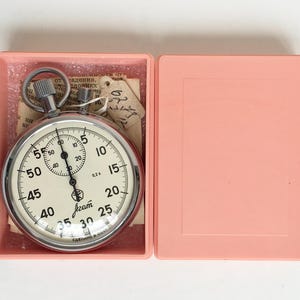 May include: A vintage stopwatch with a silver-colored case and a white face, displayed in an open pink plastic box. The stopwatch features black numerals and markings, with a smaller dial at the top. The box is open, revealing the stopwatch and some paper.