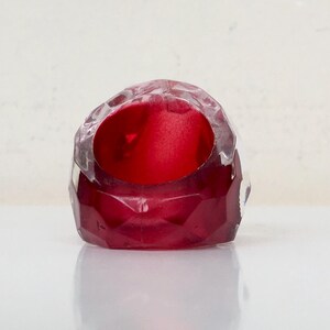 May include: A red glass ring with a faceted design. The ring is clear on the top and bottom, with a red center.