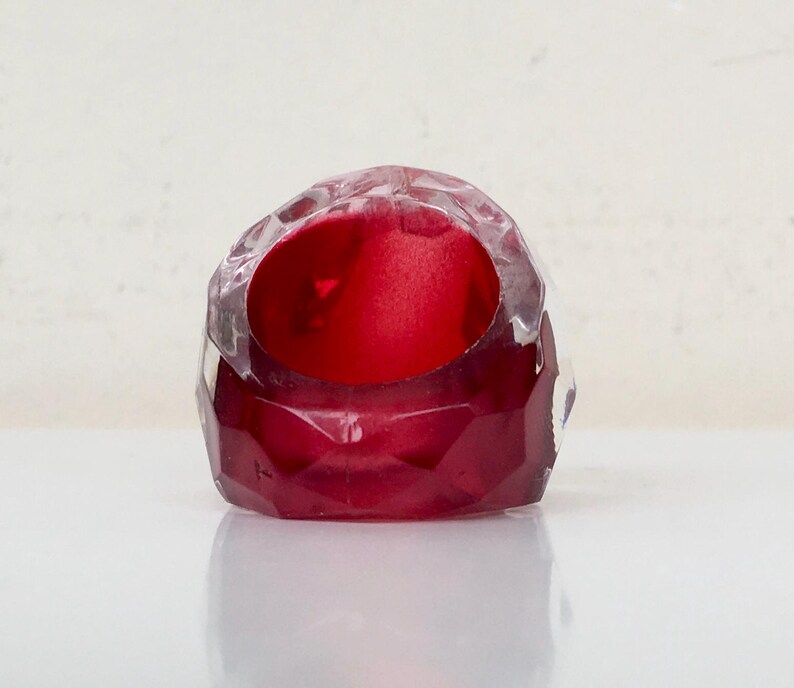 May include: A red glass ring with a faceted design. The ring is clear on the top and bottom, with a red center.