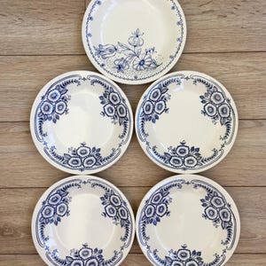 May include: A set of six white ceramic plates with a blue floral design. The plates have a scalloped edge and a detailed pattern of flowers and leaves. The plates are arranged on a wooden surface.