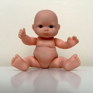 May include: A small, nude-colored baby doll with black eyes and a chubby face. The doll is sitting with its legs spread out and its arms outstretched.