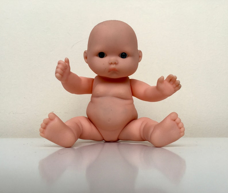 May include: A small, nude-colored baby doll with black eyes and a chubby face. The doll is sitting with its legs spread out and its arms outstretched.