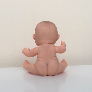 May include: A small, nude-colored baby doll sitting with its legs crossed. The doll has a realistic face and is made of soft plastic.