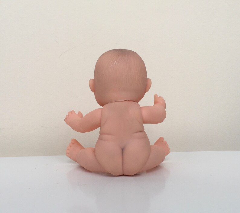 May include: A small, nude-colored baby doll sitting with its legs crossed. The doll has a realistic face and is made of soft plastic.
