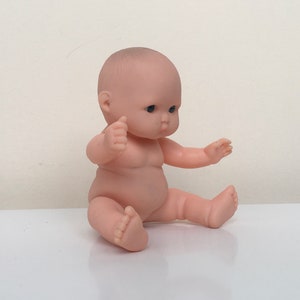 May include: A small, nude-colored baby doll with blue eyes sits with its legs crossed and arms outstretched. The doll is made of plastic and has a realistic appearance.