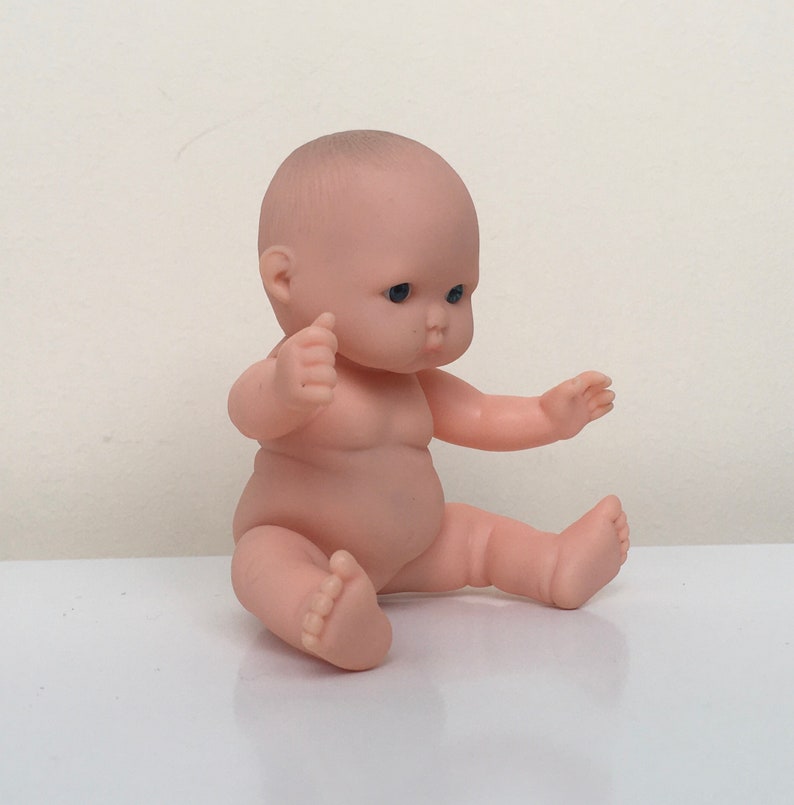 May include: A small, nude-colored baby doll with blue eyes sits with its legs crossed and arms outstretched. The doll is made of plastic and has a realistic appearance.