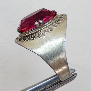 May include: A silver ring with a large, faceted, dark pink gemstone set in a decorative bezel. The ring has a wide band with a textured design.