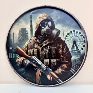 Könnte beinhalten: Ein rundes Metallschild mit einer Figur in einer getarnten Kapuzenjacke und einer Gasmaske, die ein Gewehr hält. Der Hintergrund zeigt eine Industrielandschaft mit einem hohen Schornstein und einem großen Riesenrad unter einem bewölkten Himmel.