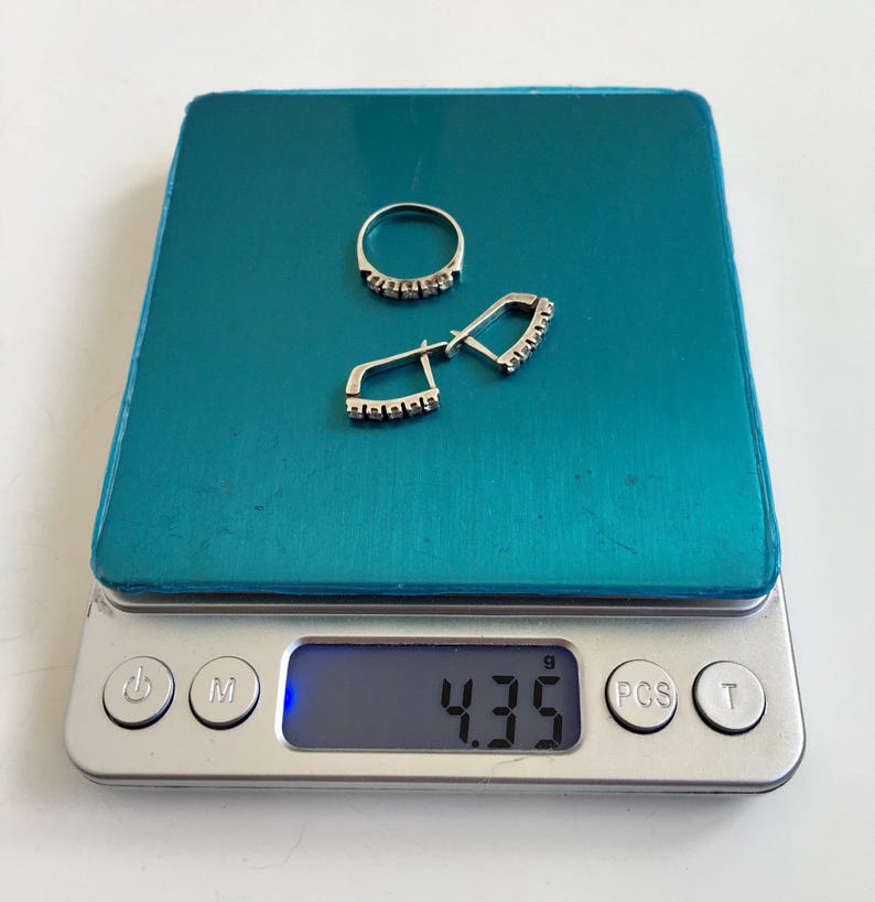 May include: A silver ring and earrings with small clear stones on a digital scale that reads 4.35 grams.