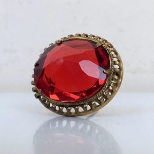 May include: A vintage ring with a large, oval, faceted red gemstone. The gemstone is set in a gold-toned metal filigree setting. The ring is displayed on a white surface.
