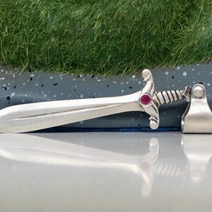 May include: A silver-colored sword pendant with a red gemstone in the hilt. The pendant has a detailed handle and a curved blade. The sword is displayed on a reflective surface, showcasing its design and craftsmanship.