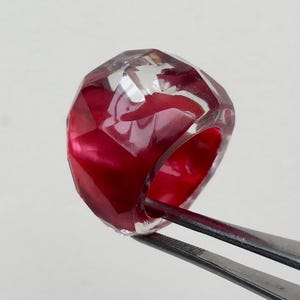 May include: A clear, faceted ring with a red interior. The ring is held by tweezers.