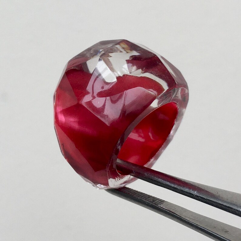 May include: A clear, faceted ring with a red interior. The ring is held by tweezers.