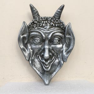 May include: A silver-colored devil face sculpture with horns, pointed ears, and a smiling expression. The sculpture has a textured surface and is likely made of metal. The devil's face is centered in the image.