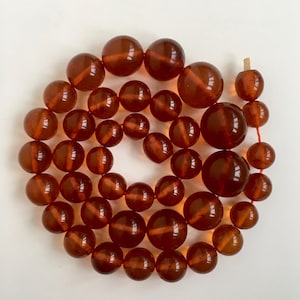 Puede incluir: Una larga hebra de cuentas de &aacute;mbar, cada cuenta es de un color marr&oacute;n intenso y rico y tiene un acabado liso y pulido. Las cuentas est&aacute;n enhebradas en un hilo rojo fino.