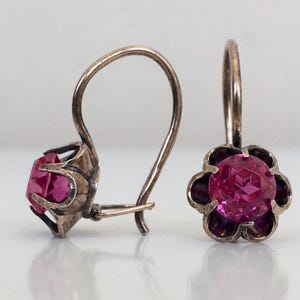 May include: A pair of silver earrings with pink gemstone accents. The earrings have a flower-shaped design with a single round gemstone in the centre.