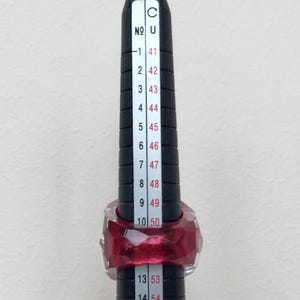 May include: A red ring with a faceted design is measured on a ring sizer. The ring sizer shows sizes from 41 to 56.
