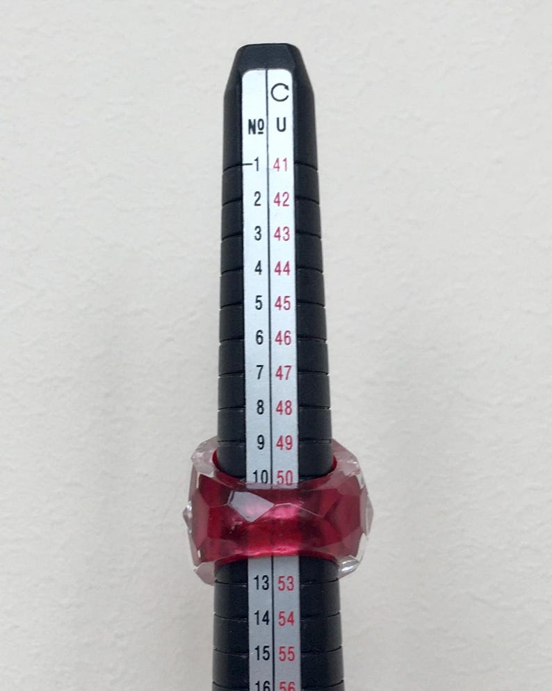 May include: A red ring with a faceted design is measured on a ring sizer. The ring sizer shows sizes from 41 to 56.