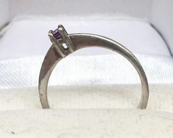 Puede incluir: Un anillo de plata con una pequeña gema púrpura de corte cuadrado. El anillo tiene un diseño sencillo y clásico, con una banda delgada. La gema es el punto focal, engastada en un engaste de cuatro puntas. El anillo se muestra en una caja blanca.