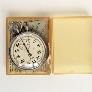 May include: A vintage stopwatch with a silver case and a cream-colored face, displayed in an open, clear plastic case. The stopwatch features black numerals and markings, with a smaller dial at the top. A small piece of paper with handwritten text is visible.