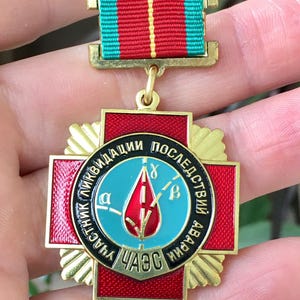 Pode incluir: Medalha dourada com um design de cruz vermelha e preta. O centro apresenta um círculo azul com uma chama vermelha e texto cirílico. Uma fita listrada verde, vermelha e amarela está presa no topo.
