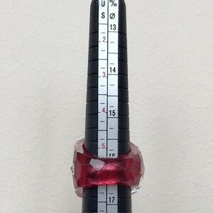 May include: A red and clear ring sized on a ring sizer with markings in millimeters and US ring sizes. The ring is a geometric shape with many facets.