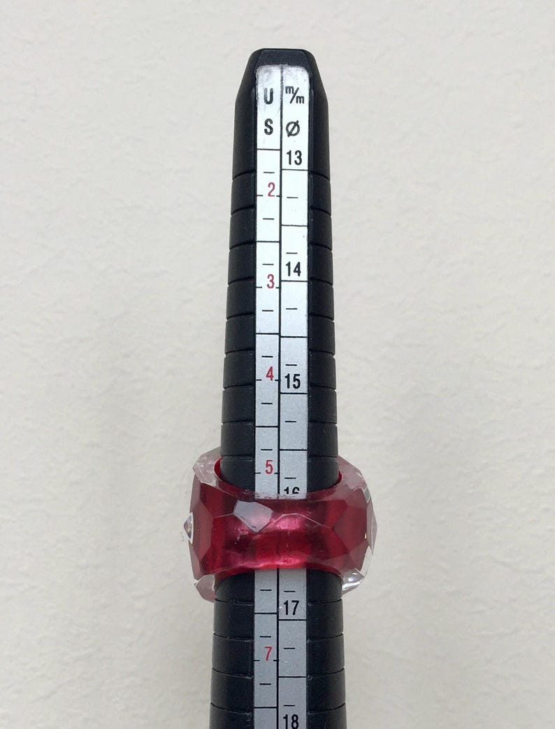 May include: A red and clear ring sized on a ring sizer with markings in millimeters and US ring sizes. The ring is a geometric shape with many facets.
