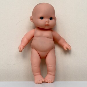May include: A small, nude-colored baby doll with black eyes and a serious expression. The doll is standing with its feet together and its arms slightly out to the sides.