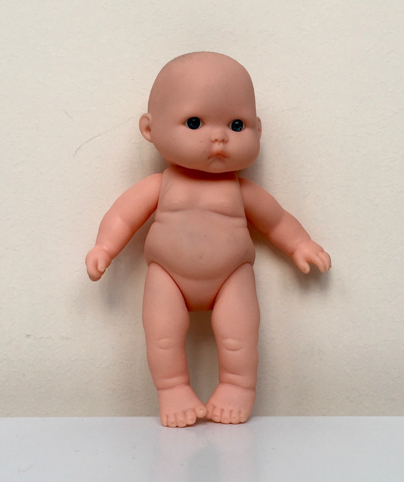 May include: A small, nude-colored baby doll with black eyes and a serious expression. The doll is standing with its feet together and its arms slightly out to the sides.