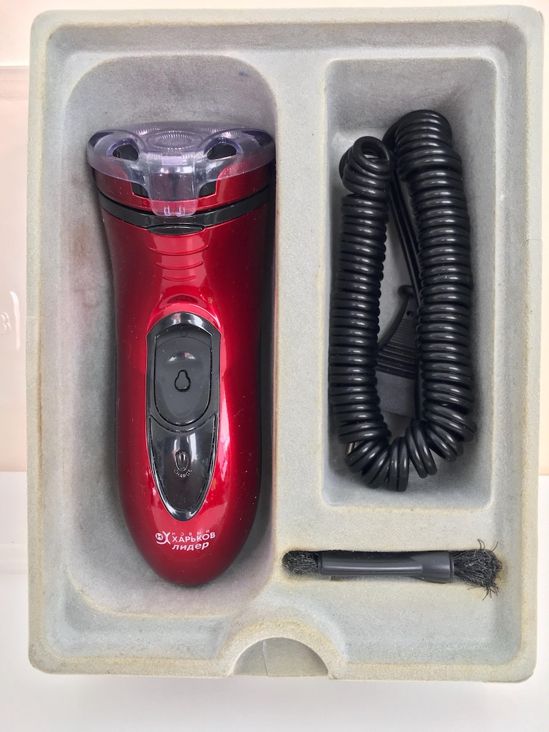 May include: A red electric shaver with a clear head and a black cord. The shaver is in a gray plastic box with a black cleaning brush.