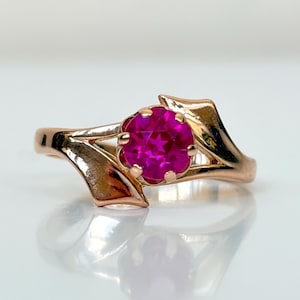 May include: A rose gold ring featuring a round, faceted, vibrant pink gemstone set in a six-prong mounting. The band has a polished finish and a unique, asymmetrical design that tapers toward the center setting, creating a sleek, metallic look.