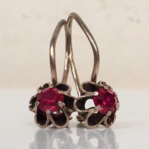 May include: A pair of silver-toned earrings with bright red, round gemstones. The gemstones are set in a flower-shaped setting with dark accents. The earrings have a simple hook design.