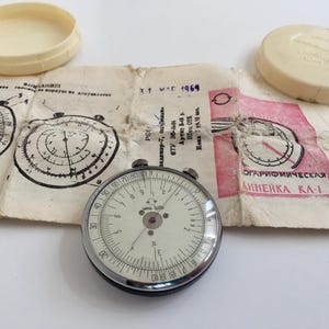 Pode incluir: Uma régua de cálculo vintage com um mostrador circular, rodeada por uma moldura de metal e dois recipientes de plástico creme. A régua de cálculo está colocada sobre um pedaço de papel envelhecido com diagramas e texto. O texto "ОГАРИФМИЧЕСКАЯ ЛИНЕЙКА КА-1" é visível.