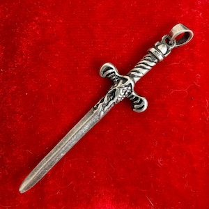 May include: A silver-toned metal pendant shaped like a sword. The hilt features intricate, textured detailing with a twisted grip and a crossguard. A small circular loop is attached to the pommel for hanging, and the blade is straight and smooth.