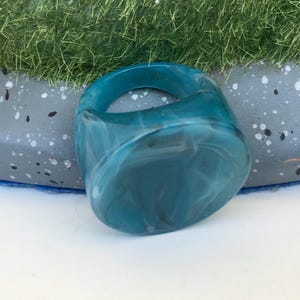 Puede incluir: Un anillo grueso de resina turquesa con efecto marmolado. El anillo tiene forma rectangular redondeada y una banda gruesa. El color turquesa tiene remolinos blancos, creando un patrón único. Una pieza de joyería audaz.