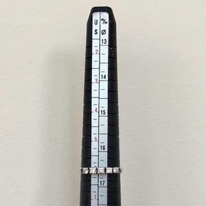 May include: A silver band ring with small diamonds set in a row. The ring is measured on a ring sizer that shows a size 17.