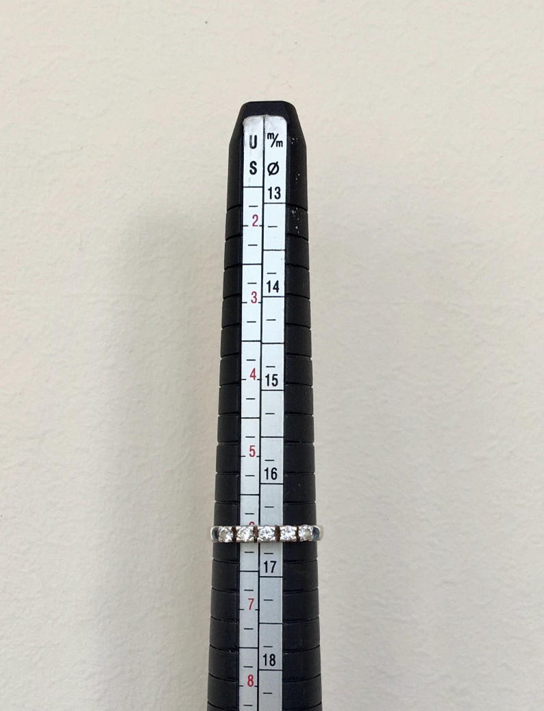 May include: A silver band ring with small diamonds set in a row. The ring is measured on a ring sizer that shows a size 17.