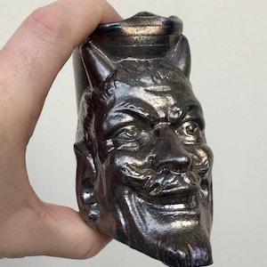 Könnte beinhalten: Eine dunkle, metallisch aussehende Keramiktasse in Form eines Teufelskopfes. Die Tasse hat Hörner, einen Schnurrbart und ein finsteres Grinsen. Die Details sind gut definiert, und der Gesamteffekt ist sowohl auffällig als auch leicht beunruhigend.
