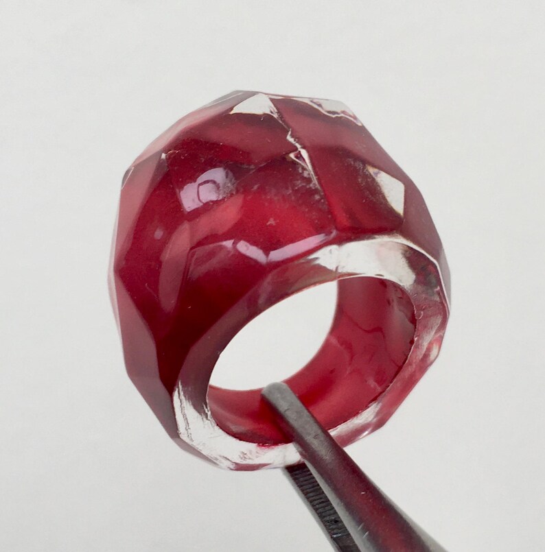 May include: A red and clear faceted ring made of resin. The ring is held by tweezers.