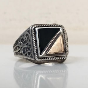 May include: A silver ring with a square face featuring a black and gold design. The ring has ornate scrollwork on the sides and a rope-like border around the face. A unique piece of jewelry.