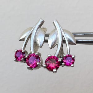 Pode incluir: Brincos de cereja prateados com pedras preciosas rosa brilhantes. Cada brinco apresenta um par de cerejas com folhas. As pedras preciosas são redondas e estão engastadas numa moldura de metal prateado. Os brincos são segurados por uma pinça.
