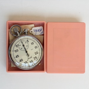 May include: A vintage stopwatch with a silver-colored casing and a cream-colored face, displayed in an open, light pink box. The stopwatch face features black numerals and markings, with the brand name "Team" visible. A small tag with the number "4669" is attached.
